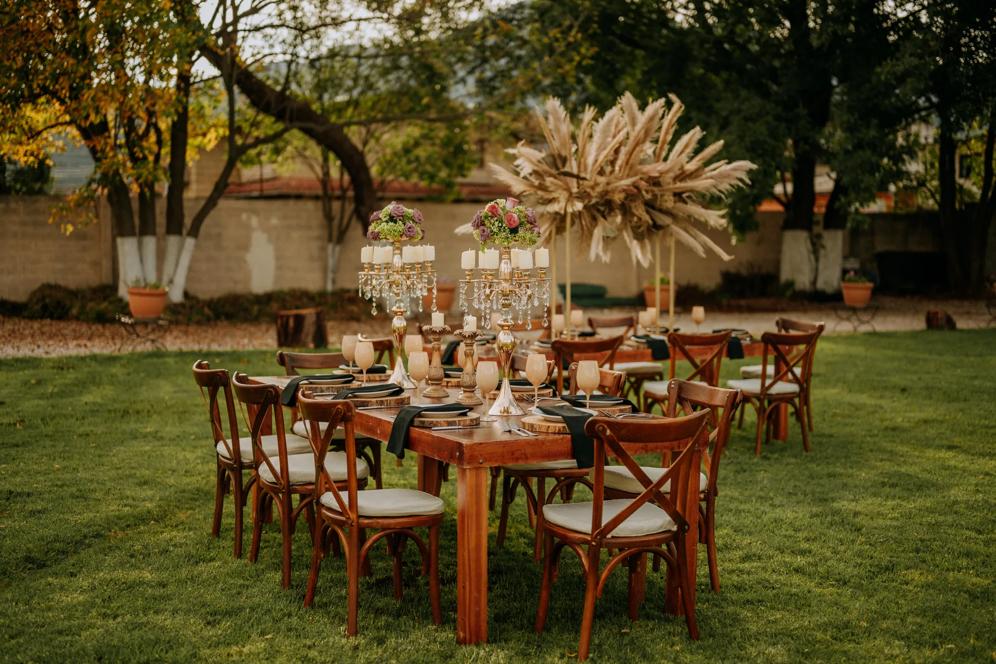 Mesa larga de madera montada en el jardín del hotel al atardecer, con candelabros y centros de pampas